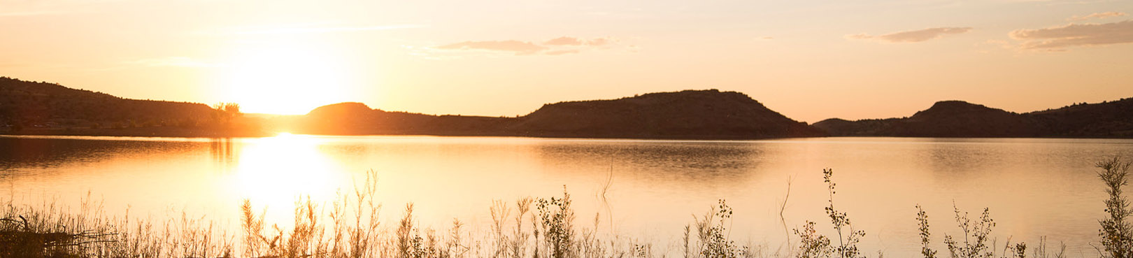 The sun sets behind the distant mountains as the orange glow reflects off the calm lake surface.